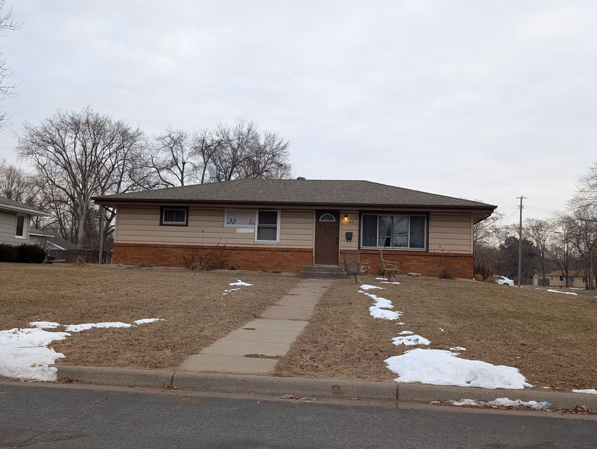 Single-story residential building with beige siding and a brick lower facade, a brown front door, and two windows. There is a concrete walkway leading up to the front door, flanked by patches of snow on the grass lawn. Two outdoor chairs are placed near the entrance. Leafless trees and a cloudy sky are visible in the background.