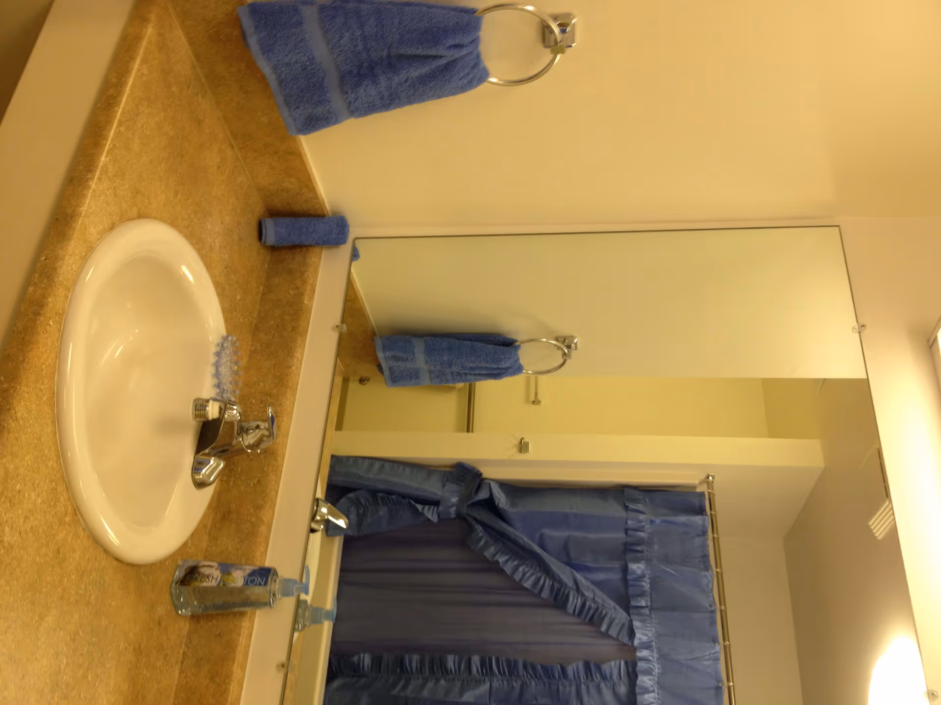 Bathroom sink with a beige countertop, a chrome faucet, a bottle of hand soap, and blue towels hanging on a ring and rolled on the counter. A large mirror reflects a blue shower curtain and part of the bathroom wall.