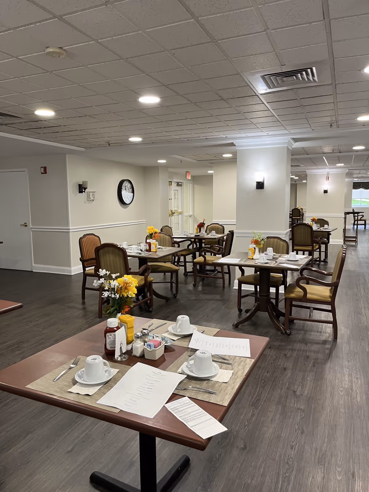 A dining room in a senior living facility with several tables set for meals. Each table has placemats, cups, utensils, menus, condiments, and small flower arrangements. The room has wood flooring, white walls with chair rails, and recessed ceiling lights. There are multiple chairs around each table and a clock on one wall.