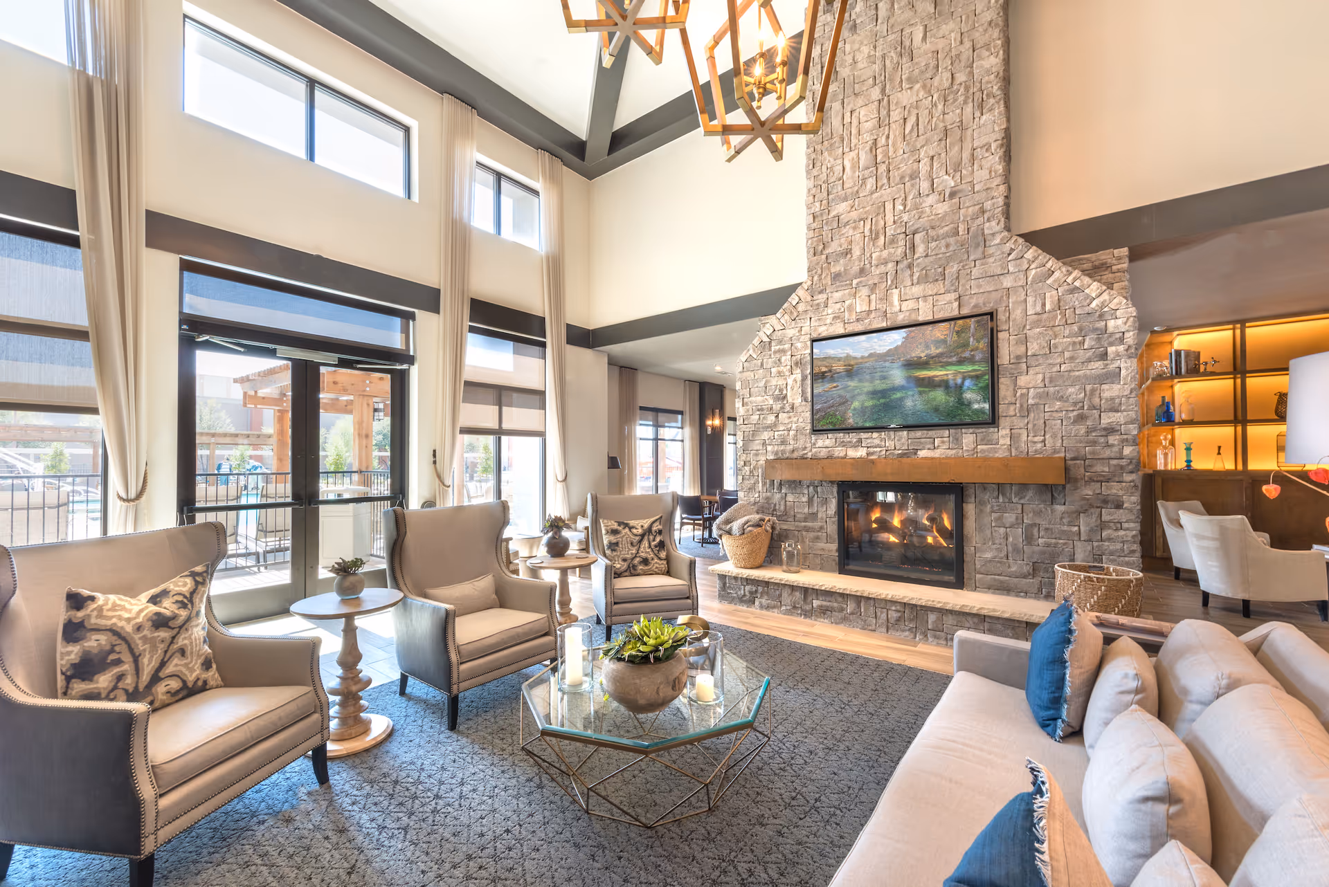 A bright and spacious living room with high ceilings and large windows. The room features a stone fireplace with a mounted TV above it, beige armchairs with patterned pillows, a glass coffee table with candles and a plant, and a beige sofa with blue and beige pillows. There are also side tables and a cozy, inviting atmosphere.