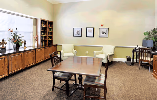 A bright communal sitting room with a wooden table and chairs, two armchairs, bookshelves, and a computer desk.