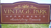 A large maroon and gold sign for Vintage Park Apartments, an assisted living community, displayed outdoors on a grassy area.