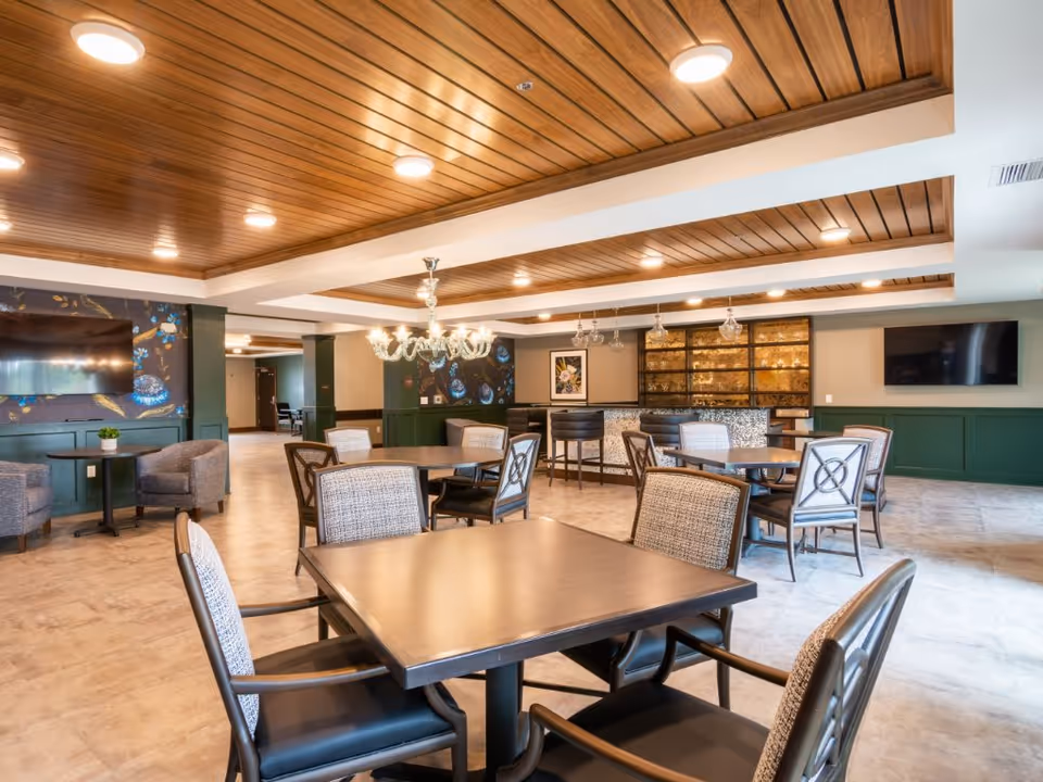 A spacious senior living facility dining room with multiple tables and chairs arranged neatly. The ceiling features wooden paneling with recessed lighting and a chandelier. There are two flat-screen TVs mounted on the walls, green wainscoting, and a bar area with high chairs and glass pendant lights. The floor is tiled with a light-colored pattern.