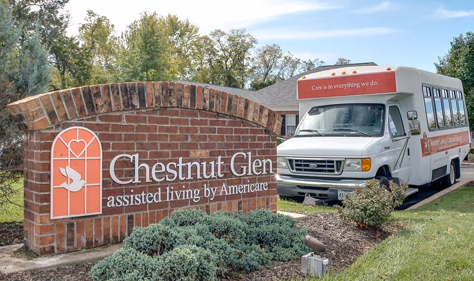 A brick sign for Chestnut Glen assisted living by Americare with a white shuttle bus parked behind it. The bus has a red banner on the front that reads 'Care is in everything we do.' There are trees and shrubs around the area under a clear sky.