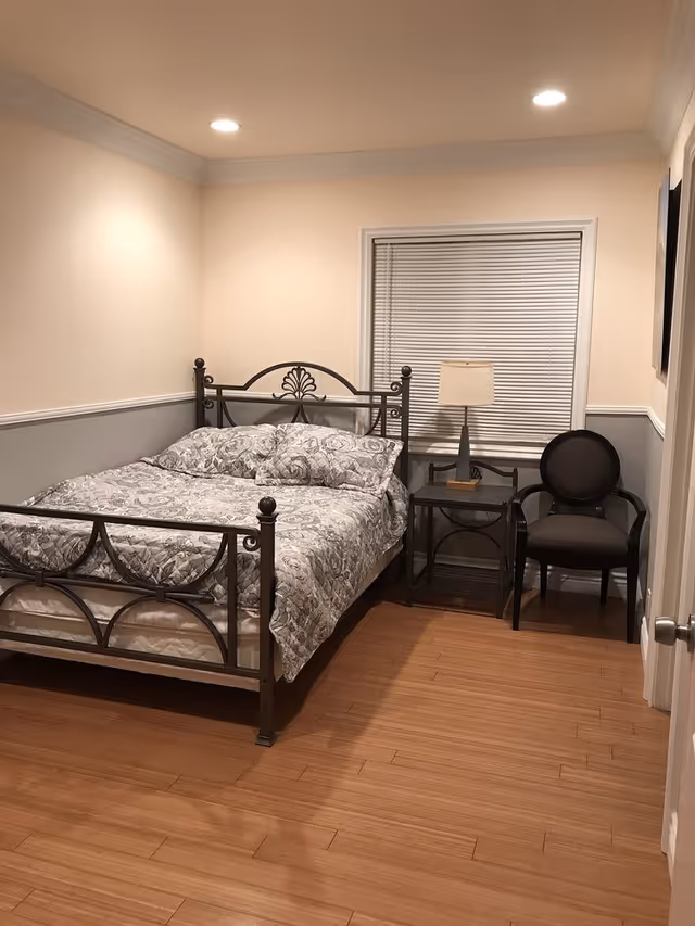 A bedroom with a metal framed bed covered in a floral patterned comforter and pillows. Next to the bed is a small black nightstand with a lamp on it, and beside the nightstand is a black armchair. The room has wooden flooring, beige walls with a gray wainscoting, a window with closed blinds, and recessed ceiling lights.
