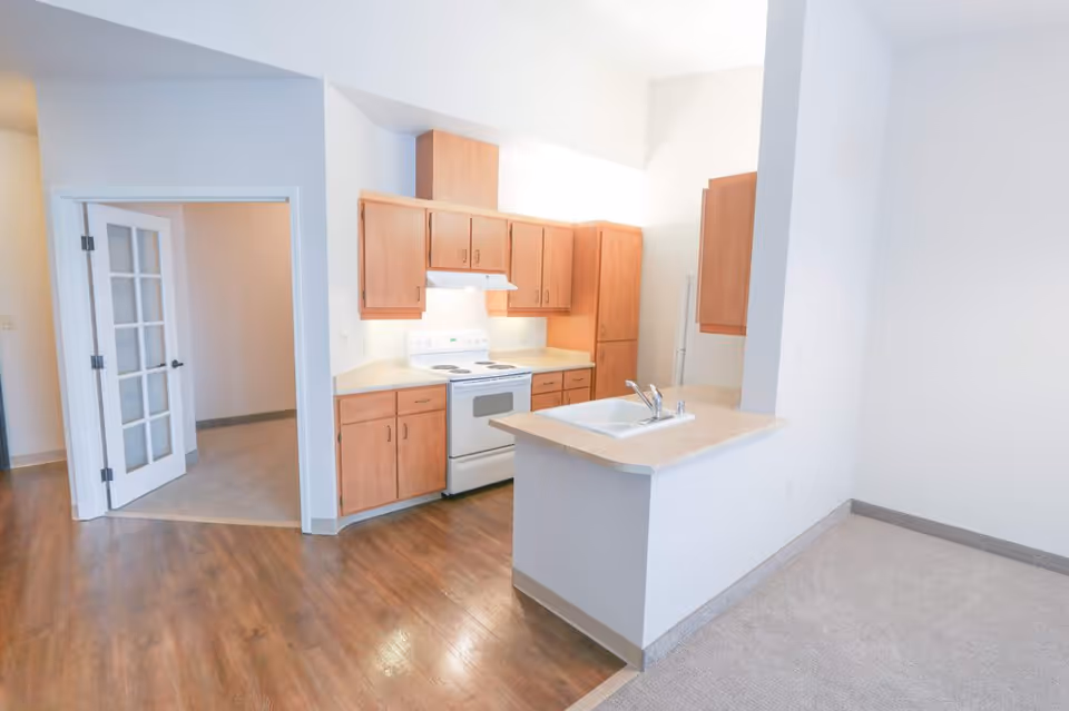 Bright kitchen area with wooden cabinets, a white stove, and a countertop with a sink. The floor is a mix of wood and carpet, and there is a doorway with glass-paneled double doors leading to another room.