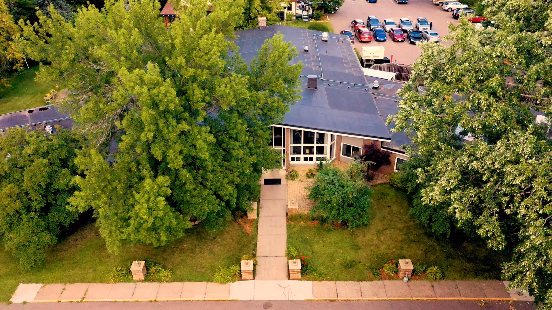 Aerial view of the front entrance of a single-story building surrounded by large green trees and grass. A concrete walkway leads to the entrance, and a parking lot with several cars is visible in the background.