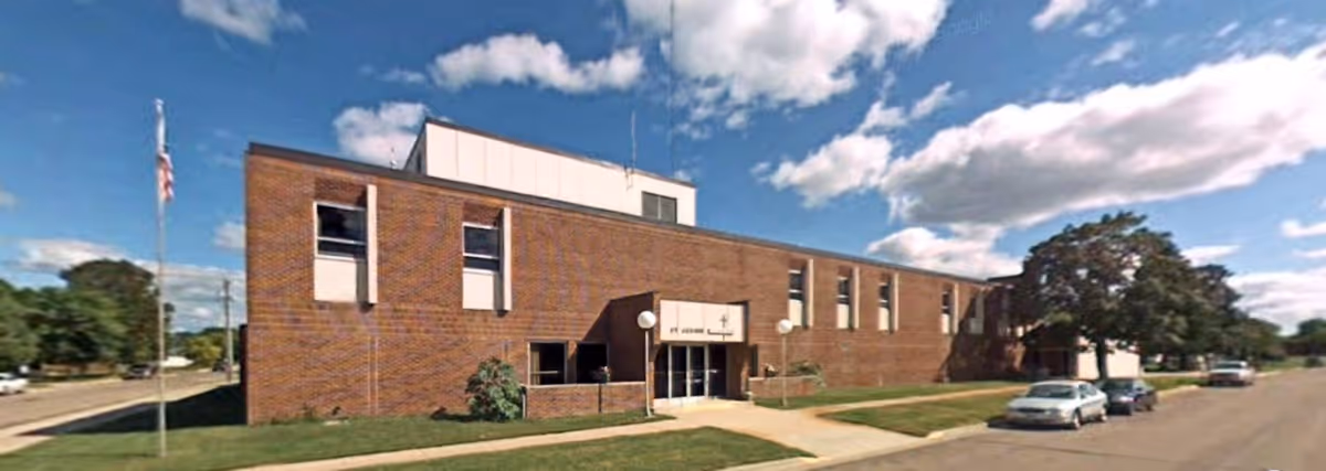 Two-story brick community building with a central entrance, flagpole, and parked cars along a residential street under a partly cloudy sky.