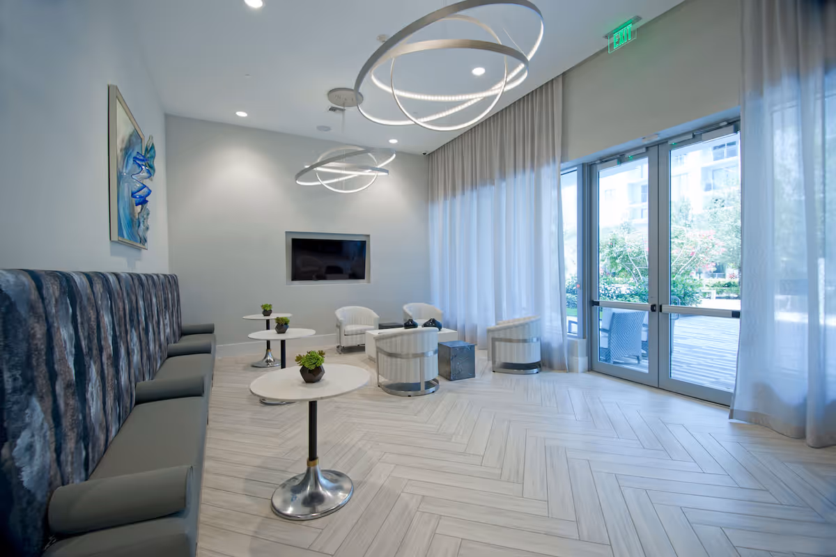 A modern lounge area with a long cushioned bench along one wall, three small round tables with potted plants, four white armchairs arranged around a small square table, large windows with sheer curtains letting in natural light, and contemporary circular ceiling lights.