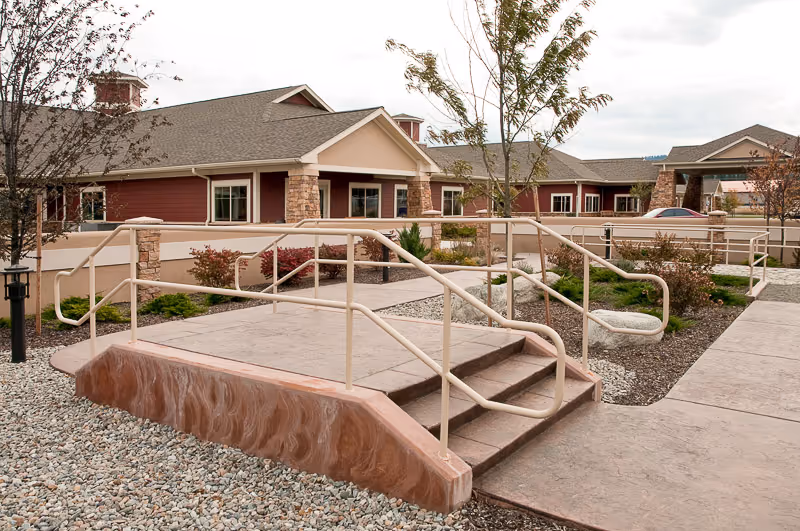 Exterior view of a senior living facility with a ramp and stairs leading to the entrance. The building has a red and beige color scheme with stone accents and a landscaped area with small trees and shrubs.
