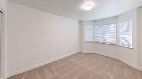 Empty carpeted room with a bay window and closed blinds, neutral walls, ceiling light, and a sliding closet door.
