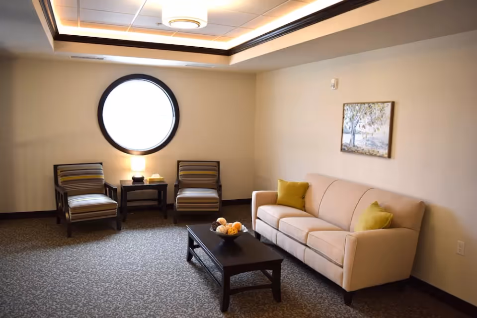 A cozy living room with a beige sofa adorned with two green pillows, two striped armchairs, a dark wooden coffee table with decorative balls, a round window, a small table with a lamp and tissues, and a painting on the wall.