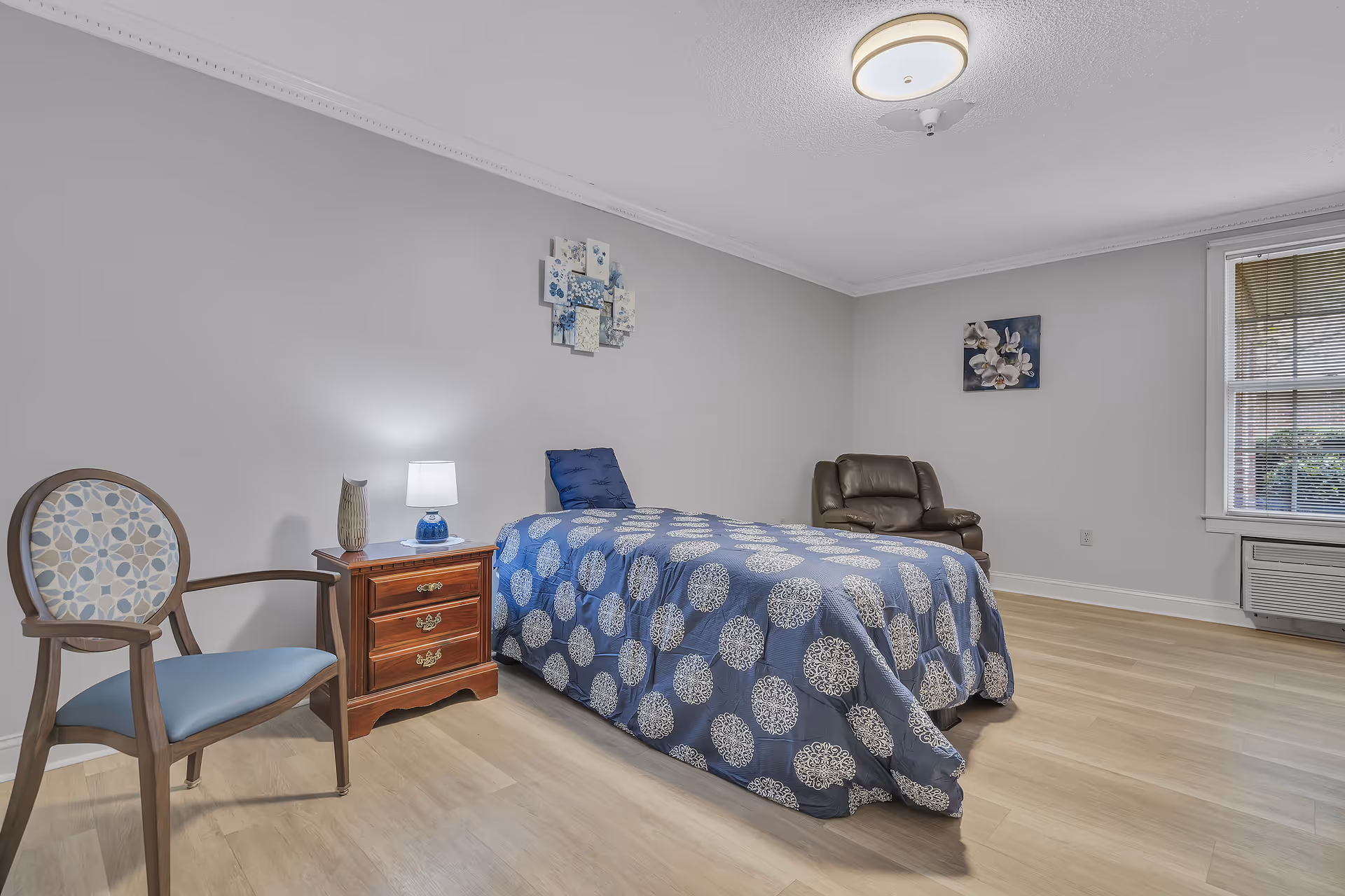 A simple bedroom with a single bed covered in a blue and white patterned bedspread. Next to the bed is a wooden nightstand with a small lamp and decorative vase. There is a wooden chair with a patterned cushion to the left of the nightstand. On the right side of the room is a brown leather armchair. The walls are light gray with two pieces of floral artwork hanging. A window with blinds is on the right wall, and the floor is light wood.