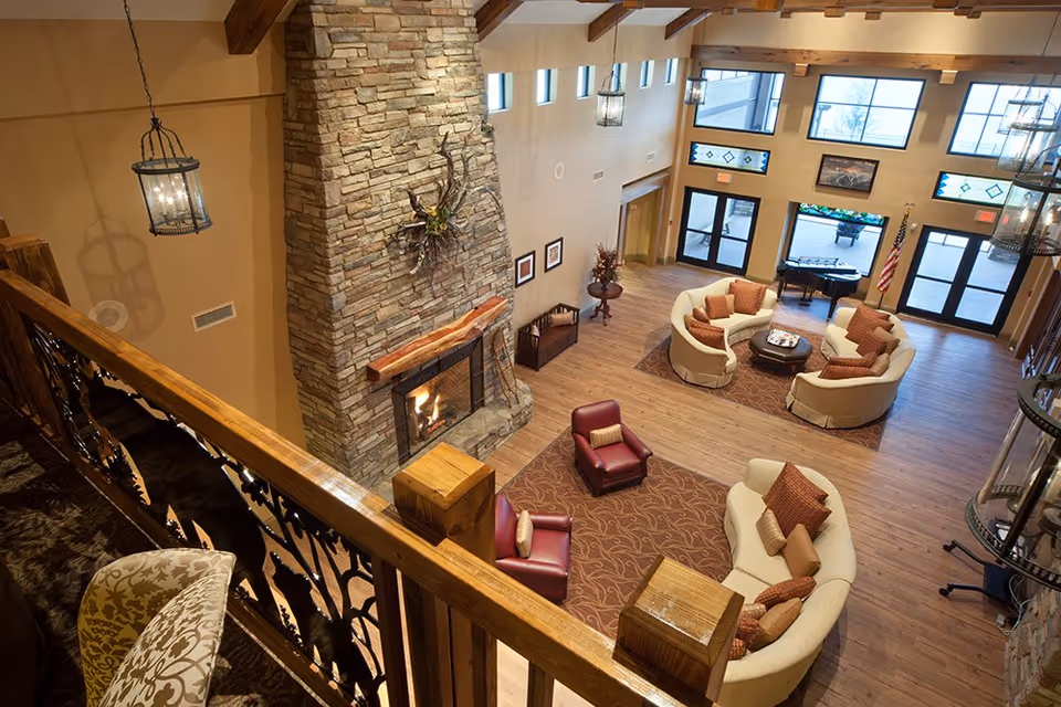 View from an upper balcony overlooking a spacious living room with a large stone fireplace, curved beige sofas with brown cushions, two red armchairs, wooden floors, and large windows letting in natural light. There is a piano near the windows and decorative lighting fixtures hanging from the ceiling.