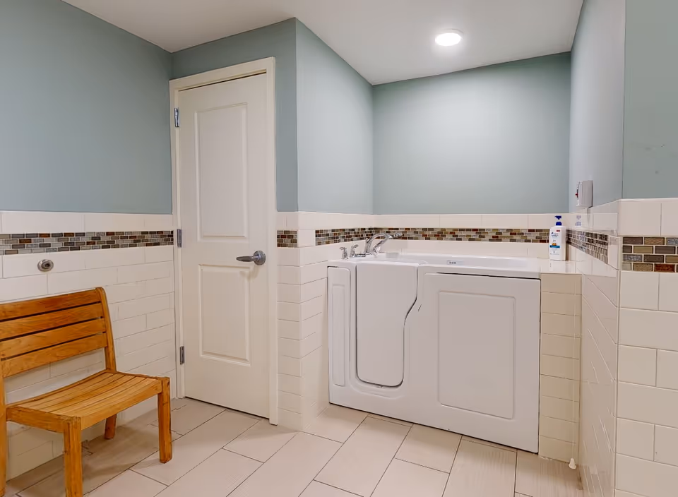 Accessible walk-in bathtub with faucets and a wooden chair in a tiled bathroom with light green walls.