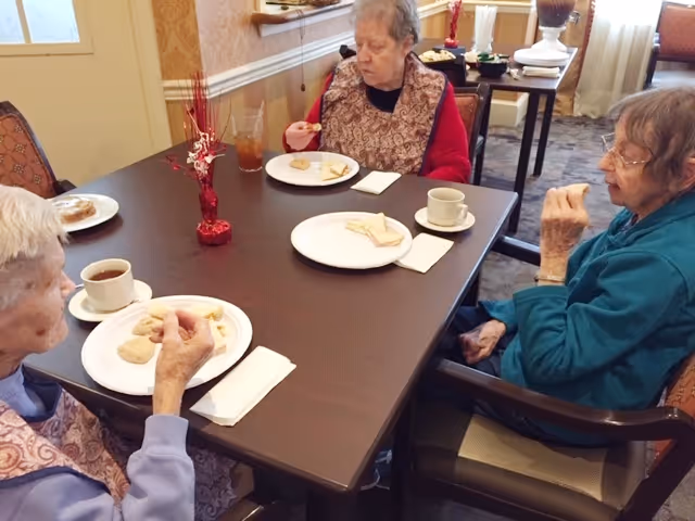 Three elderly women sitting around a dining table in a senior living facility, eating cookies and drinking coffee or tea. The table has a small red decorative centerpiece and napkins. The room has patterned carpet and light-colored walls with a window in the background.