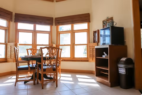 A bright dining area with a round wooden table and four matching chairs placed near large windows with brown blinds. To the right, there is a wooden shelving unit with a microwave on top and a trash bin labeled 'Aluminum Cans Only'.