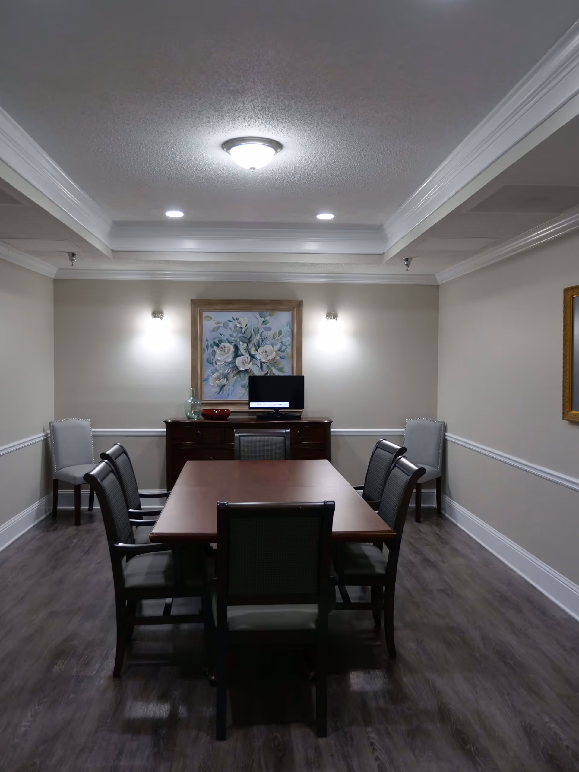 A small conference or meeting room with a rectangular wooden table surrounded by six chairs. The room has beige walls with white trim, a dark wood floor, and a ceiling light fixture with recessed lighting. On the far wall, there is a wooden sideboard with a small TV, a decorative glass vase, and a red bowl. Above the sideboard hangs a framed painting of white flowers.