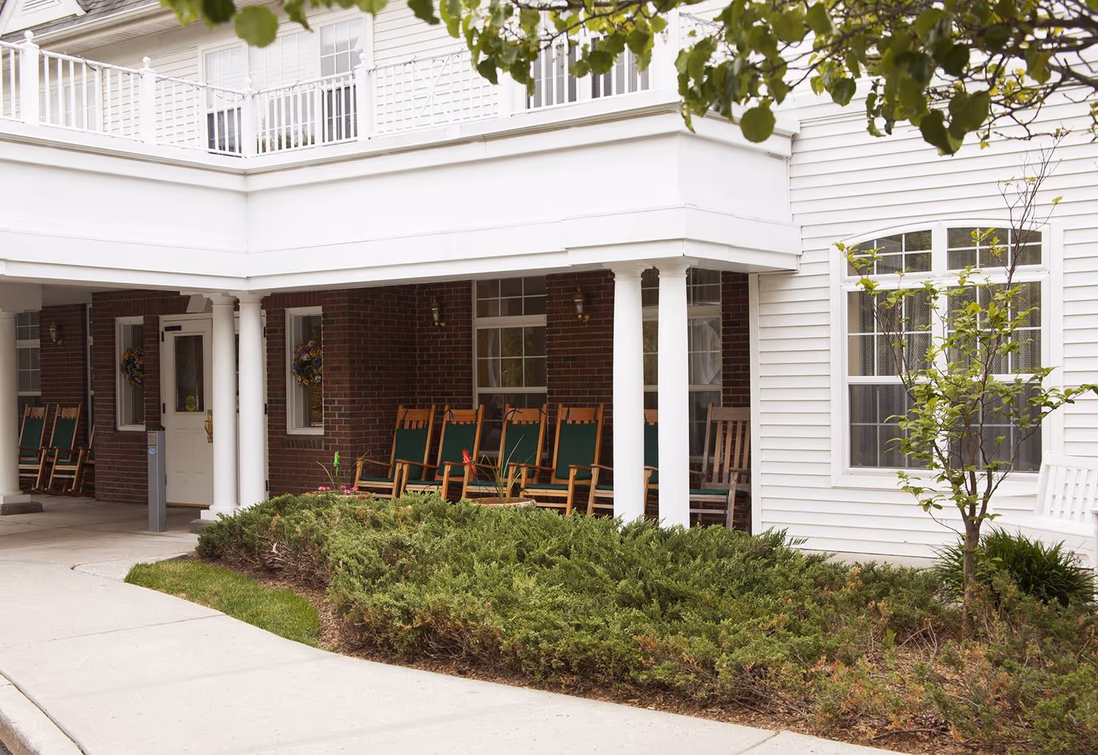 Exterior view of Brighton Gardens of Saddle River showing a covered porch area with several wooden rocking chairs with green cushions. The building features white siding and brick walls, with a small landscaped area of shrubs and a young tree in front.