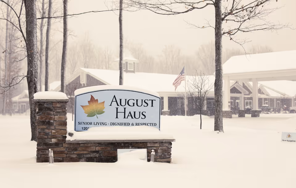 Snow-covered entrance sign reading "August Haus" in front of the senior living building with trees and an American flag.