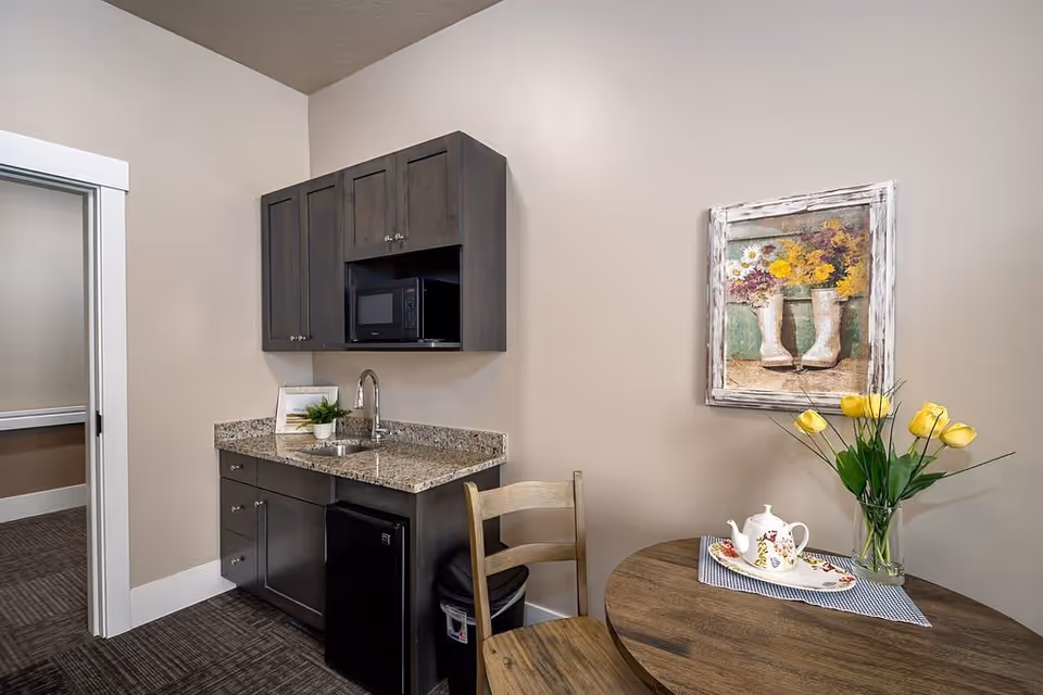 A small kitchenette area with dark wood cabinets, a granite countertop with a sink, a microwave, and a mini refrigerator. Next to the kitchenette is a round wooden table with a vase of yellow tulips and a teapot on a tray. A framed painting of flowers in white boots hangs on the beige wall above the table.