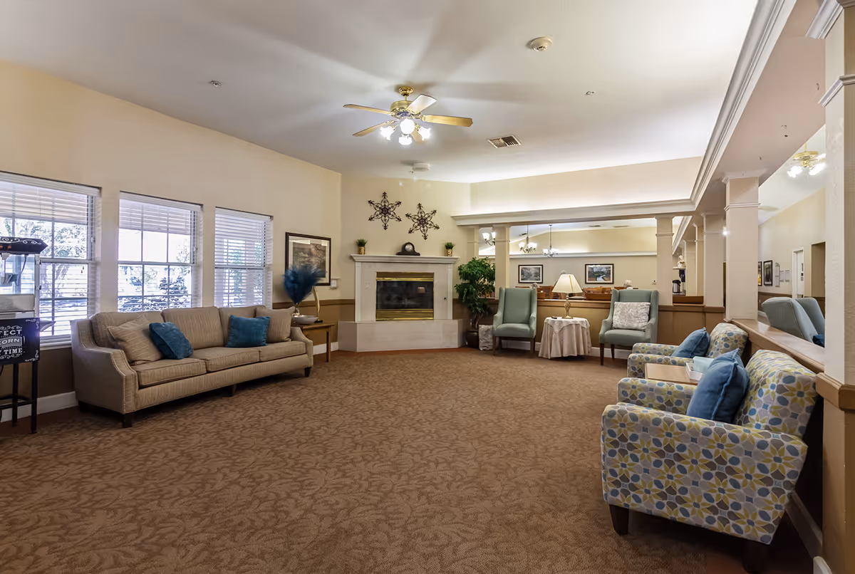 A spacious senior living facility common area with beige walls and carpet. The room features a fireplace with decorative wall art above it, a ceiling fan with lights, and several seating options including a beige sofa with blue cushions, two patterned armchairs with blue cushions, and two green armchairs around a small table with a lamp. Large windows allow natural light to fill the room.