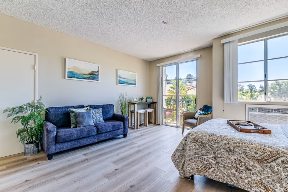 A bright and spacious senior living bedroom featuring a patterned bedspread, a blue sofa with decorative pillows, two framed landscape paintings on the wall, a small table with two stools near a sliding glass door leading to a balcony, a wicker chair, and large windows letting in natural light.