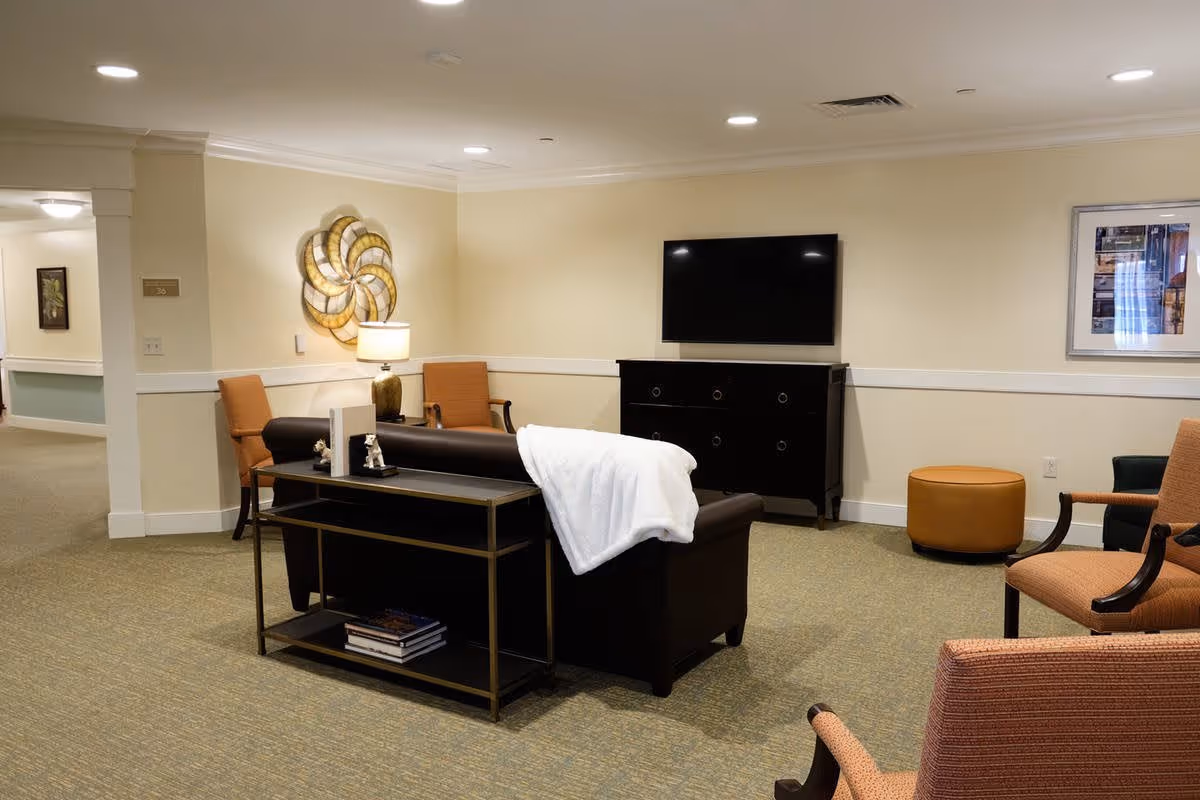 A cozy senior living facility common area with a dark brown leather sofa draped with a white blanket, two orange armchairs, a small round ottoman, a black TV stand with a flat-screen TV mounted on the wall above it, a decorative wall piece, and a lamp on a side table. The room has beige walls and carpeted flooring.