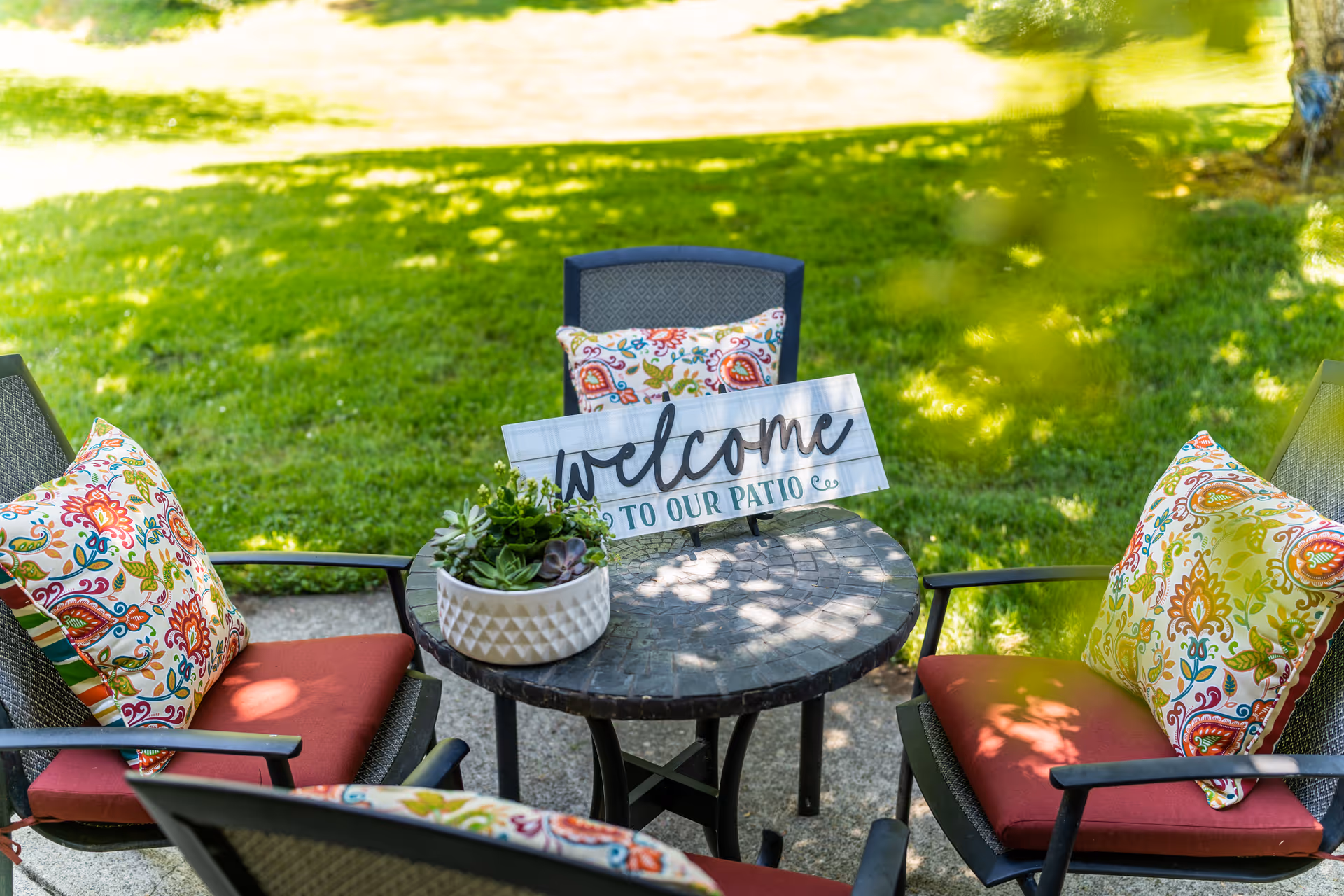 A round outdoor patio table with a small potted succulent plant and a decorative sign that reads 'welcome TO OUR PATIO' surrounded by four chairs with red cushions and colorful patterned pillows, set on a concrete patio with green grass and trees in the background.