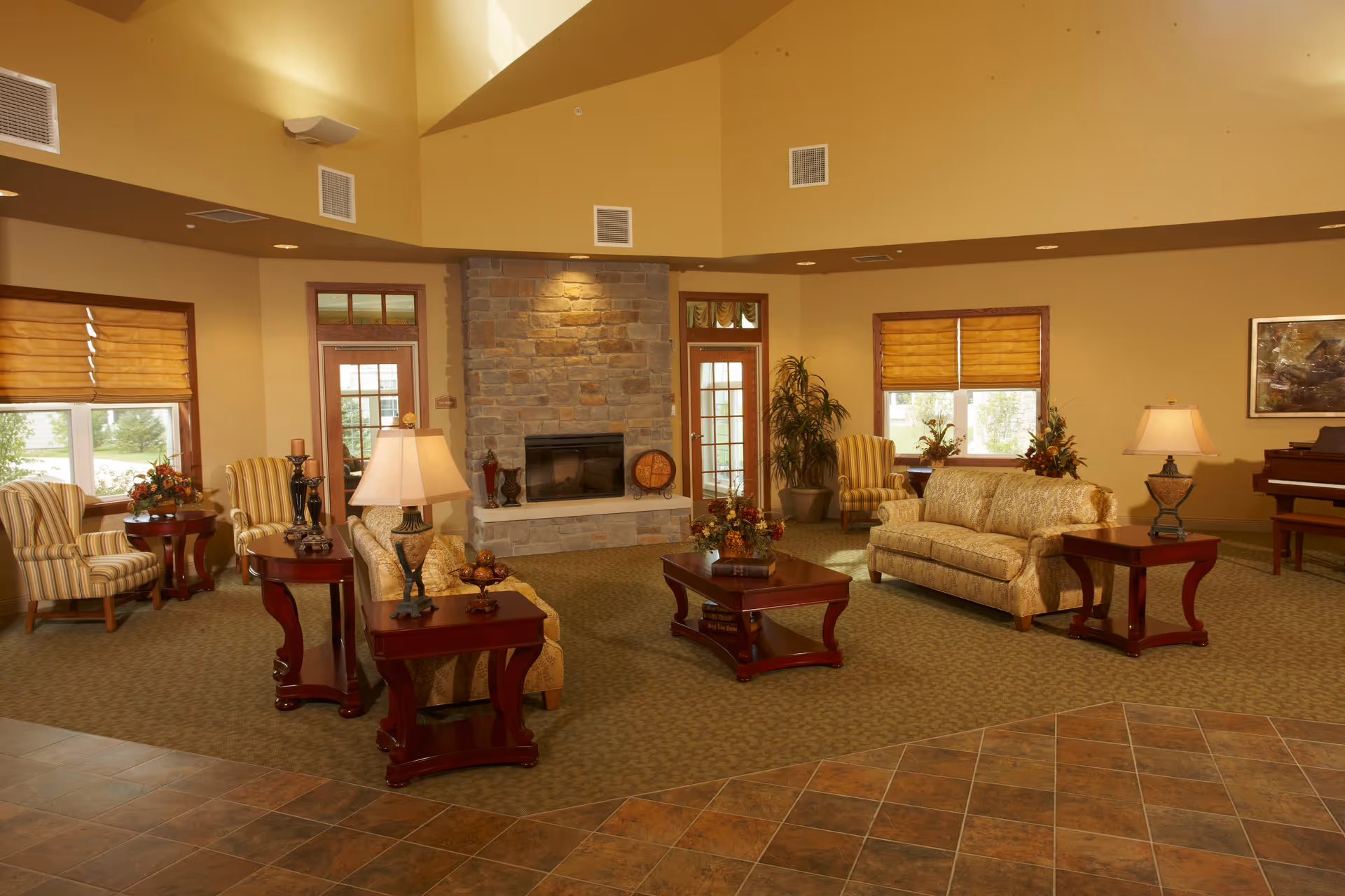 A spacious living room with beige walls and carpet, featuring a stone fireplace in the center. The room is furnished with patterned sofas, striped armchairs, wooden coffee and side tables with lamps and decorative items. Large windows with yellow blinds let in natural light, and a piano is visible in the background.
