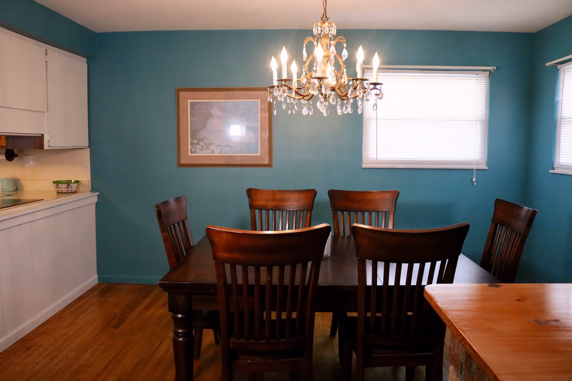A dining room with teal walls, a wooden dining table surrounded by six wooden chairs, a chandelier hanging above the table, two windows with blinds, and a framed picture on the wall.