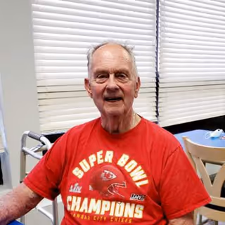 An elderly man wearing a red Kansas City Chiefs Super Bowl Champions t-shirt is sitting indoors near a window with closed blinds. A walker and a table with chairs are visible in the background.