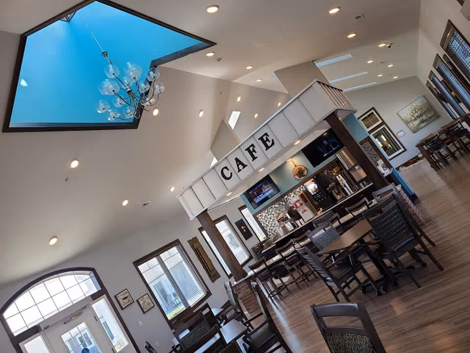 Bright open cafe-style dining area with tables and chairs, a service counter labeled "CAFE", and a skylight with a chandelier.