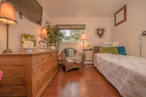 A cozy senior living bedroom with a bed, dresser, chair, plants, and warm lamps.