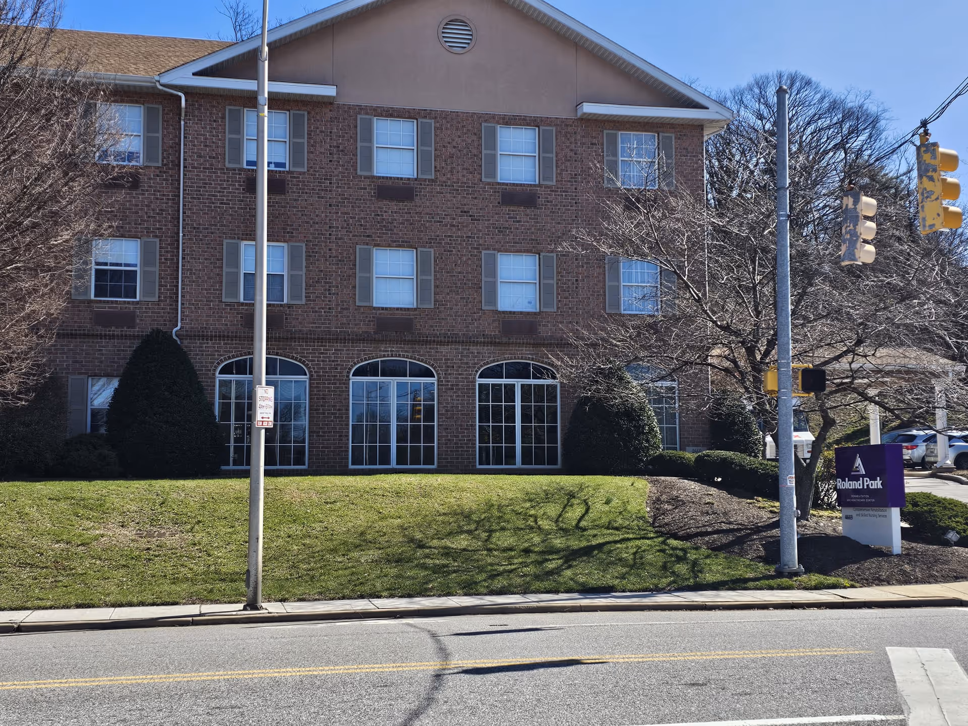 Front exterior of a three-story brick building with arched ground-floor windows, a grassy lawn and a 'Roland Park' sign near the entrance.