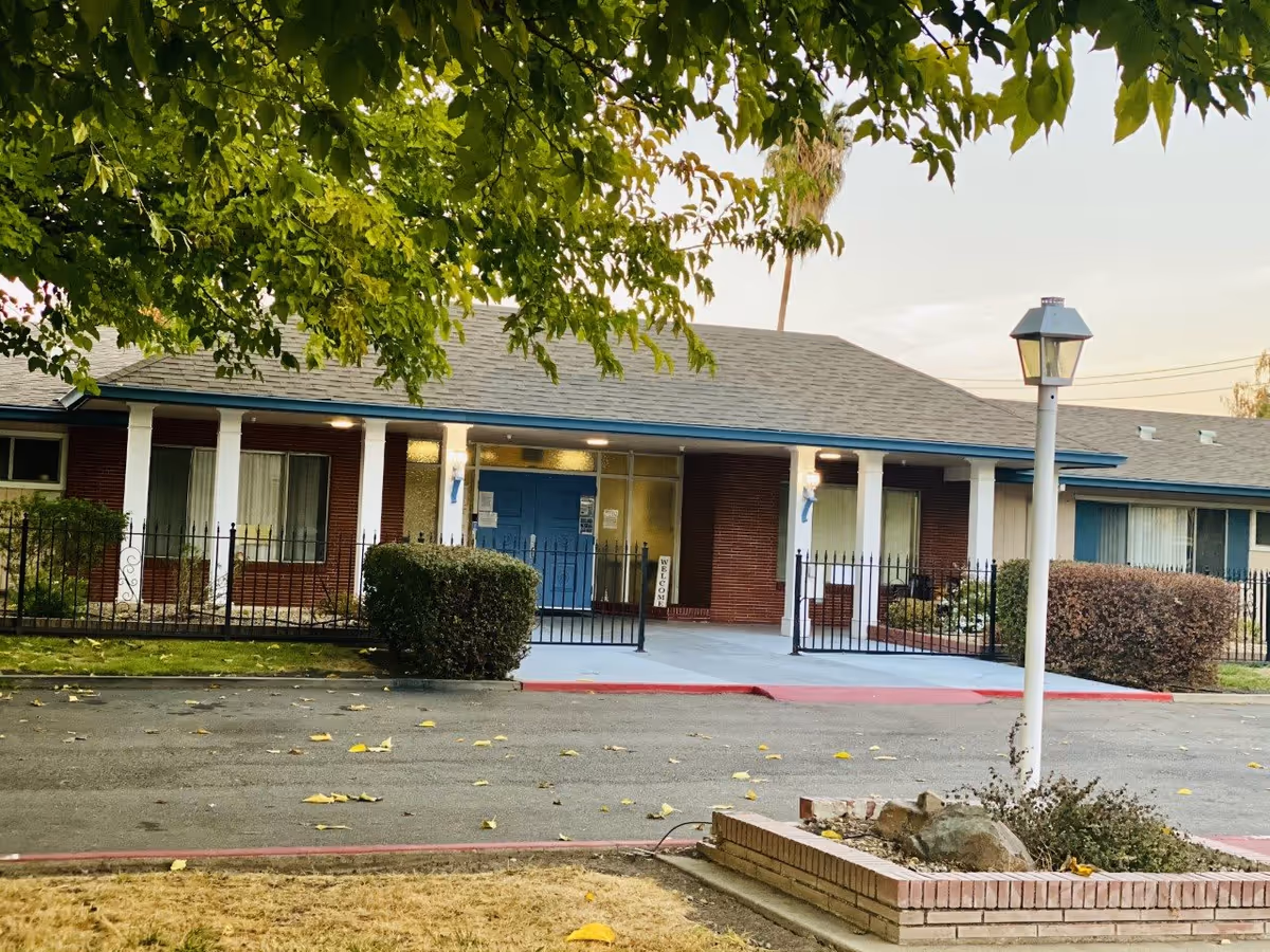 Exterior view of a single-story brick building with a blue door entrance, white pillars, and windows with curtains. There is a black metal fence in front, a lamp post on the right, and trees with green leaves partially covering the top of the image. The ground has some fallen leaves and a small garden area with rocks and plants.