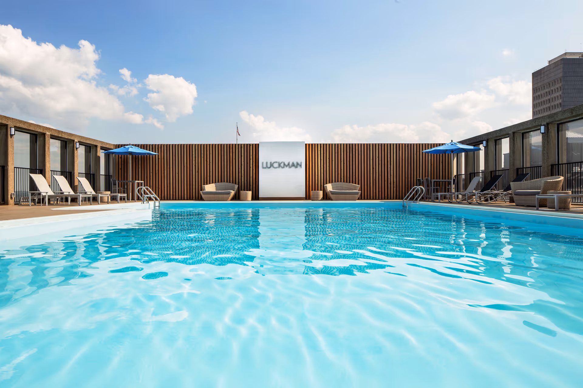 Outdoor rooftop swimming pool with clear blue water, surrounded by lounge chairs, tables with blue umbrellas, and wicker seating. A wooden fence with a white panel displaying the word 'LUCKMAN' is visible at the far end under a partly cloudy sky.