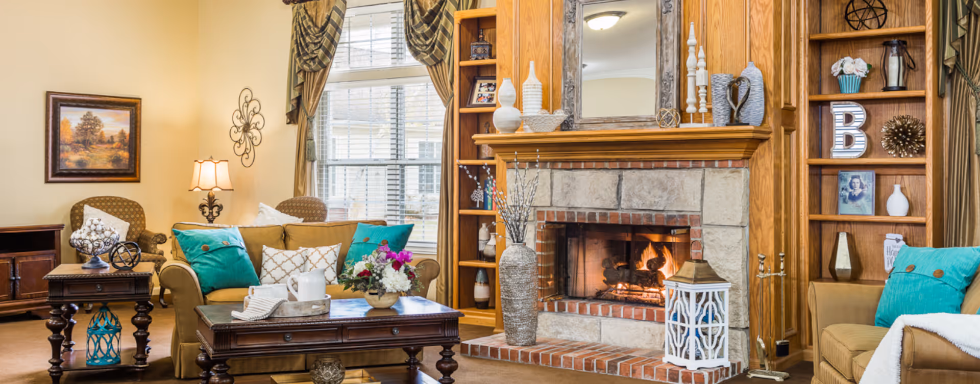 A cozy living room with a lit stone fireplace surrounded by wooden built-in shelves decorated with vases, framed photos, and decorative items. There are two armchairs and a sofa with colorful teal and white pillows, a wooden coffee table with a flower arrangement, and a window with patterned curtains letting in natural light.