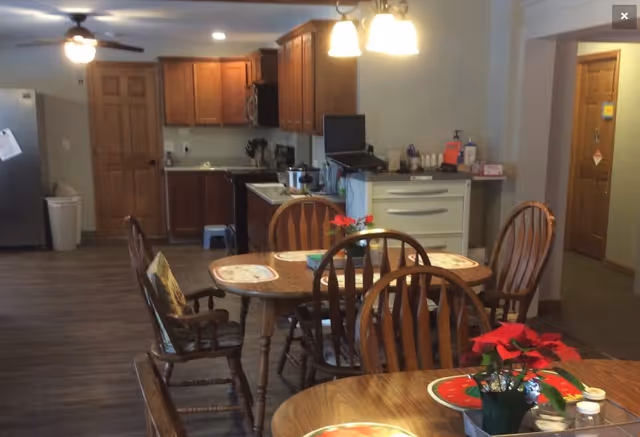 Interior view of a senior living facility dining area and kitchen. The image shows wooden dining tables with chairs, placemats, and small potted plants on the tables. In the background, there is a kitchen with wooden cabinets, a refrigerator, a stove, and a countertop with various items. The floor is wooden, and there are ceiling lights and a ceiling fan visible.