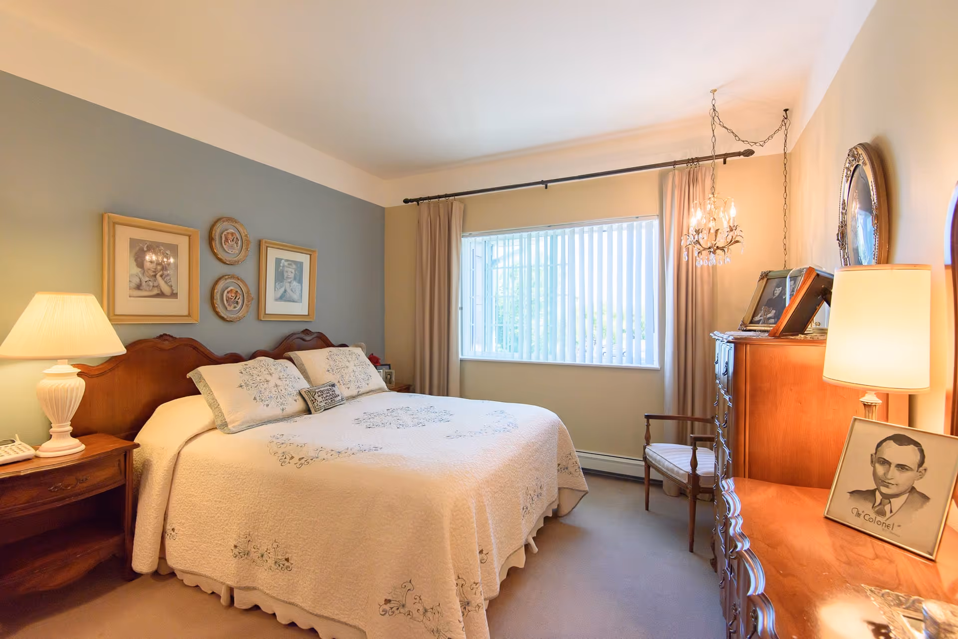 A cozy bedroom with a large bed covered in a white quilt with floral embroidery. The bed has a wooden headboard and is flanked by a wooden nightstand with a lamp and a telephone. Above the bed are framed pictures and decorative plates on a blue accent wall. A window with vertical blinds and beige curtains lets in natural light. On the right side of the room, there is a wooden dresser with framed photos and a lamp, and a wooden chair next to it. A small chandelier hangs from the ceiling near the window.