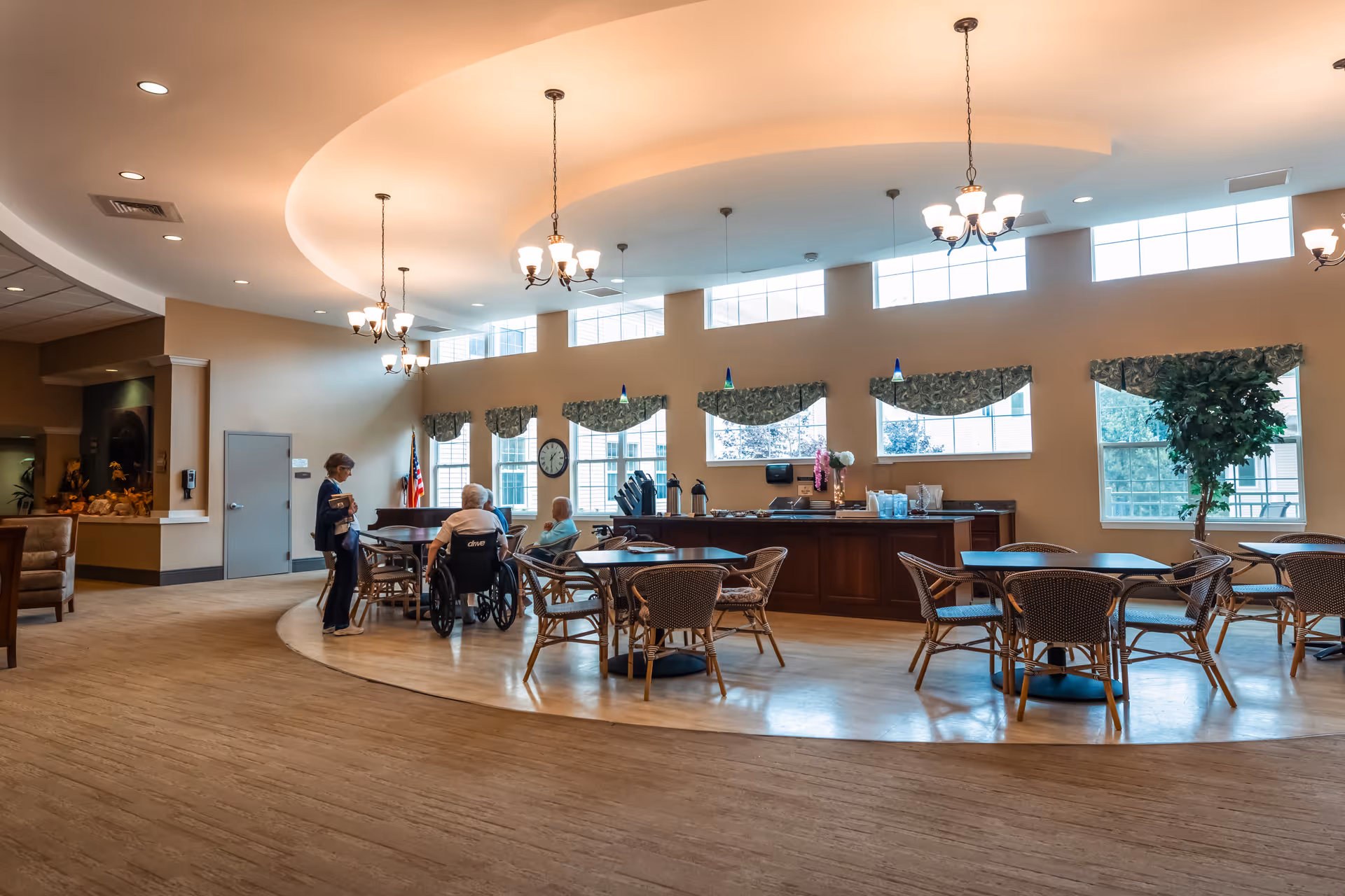 Bright dining and lounge area with round tables and chairs, a service counter, chandeliers, and seniors seated near the center.
