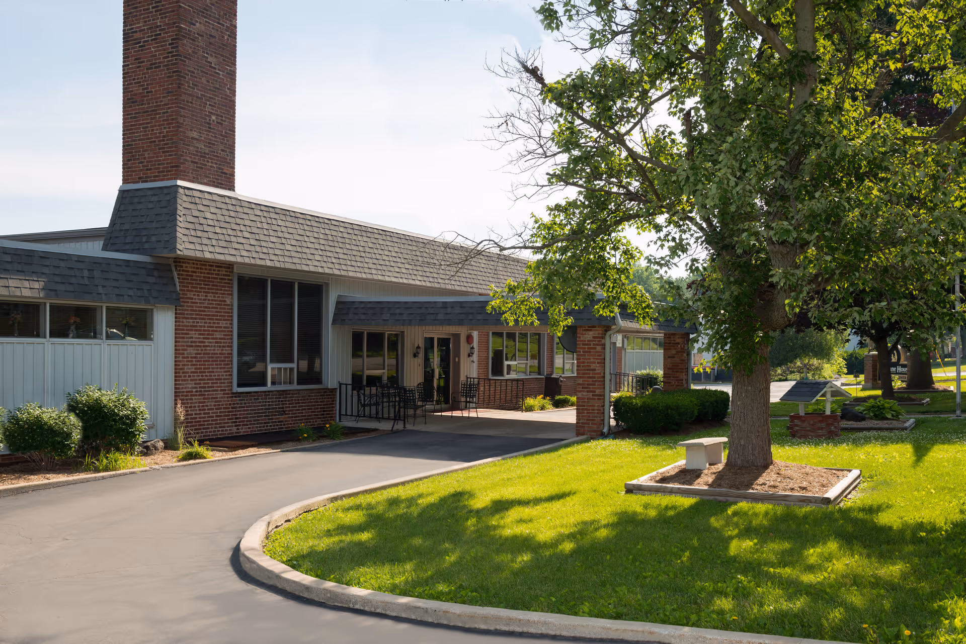 Exterior view of The Lane House facility showing a brick building with a large chimney, a covered entrance with outdoor seating, a paved driveway, and a well-maintained lawn with trees and landscaping.
