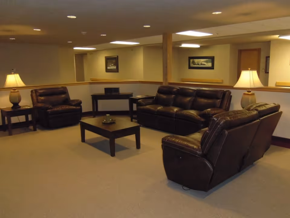 A cozy living room area with brown leather sofas and armchairs arranged around a wooden coffee table. The room is softly lit by two table lamps on side tables, with framed artwork on the walls and a beige carpeted floor.