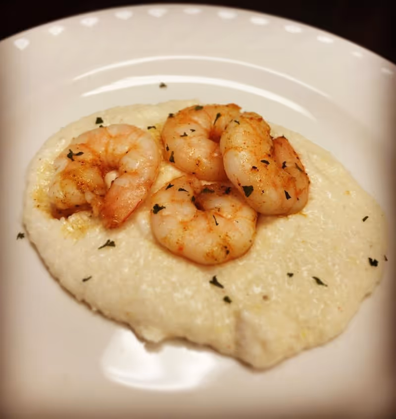 A white plate with a serving of creamy grits topped with four cooked shrimp garnished with herbs.