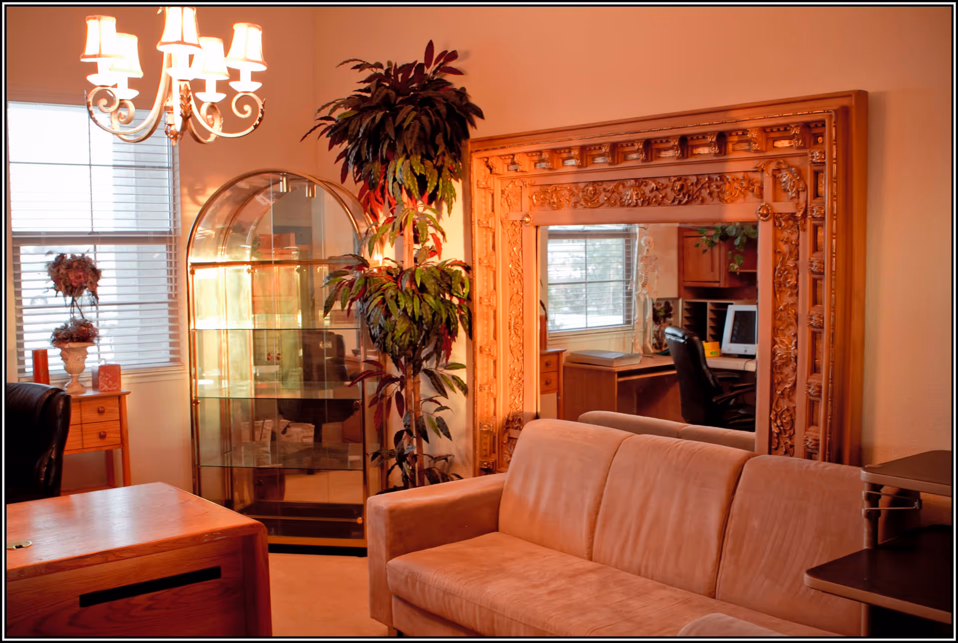 A cozy living room with a beige sofa, a large ornate wooden framed mirror reflecting a desk with a computer and office chair, a tall potted plant, a glass display cabinet, a wooden side table with a floral arrangement, and a chandelier hanging from the ceiling.
