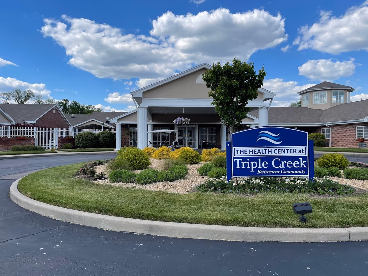 Front exterior view of The Health Center at Triple Creek Retirement Community showing a circular driveway with landscaped greenery and a blue sign with the facility's name. The building has a covered entrance with white columns and a partly cloudy blue sky above.