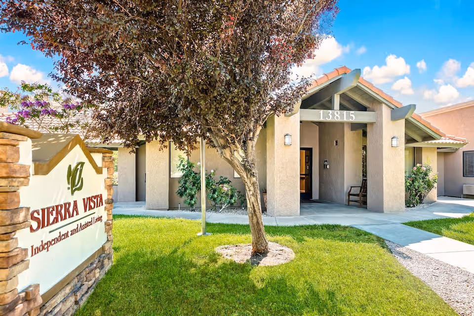 Exterior view of Sierra Vista Independent and Assisted Living facility showing the entrance with the address number 13815 above the doorway, a tree in the foreground, and a sign with the facility name on a stone base surrounded by green grass and plants under a blue sky with clouds.
