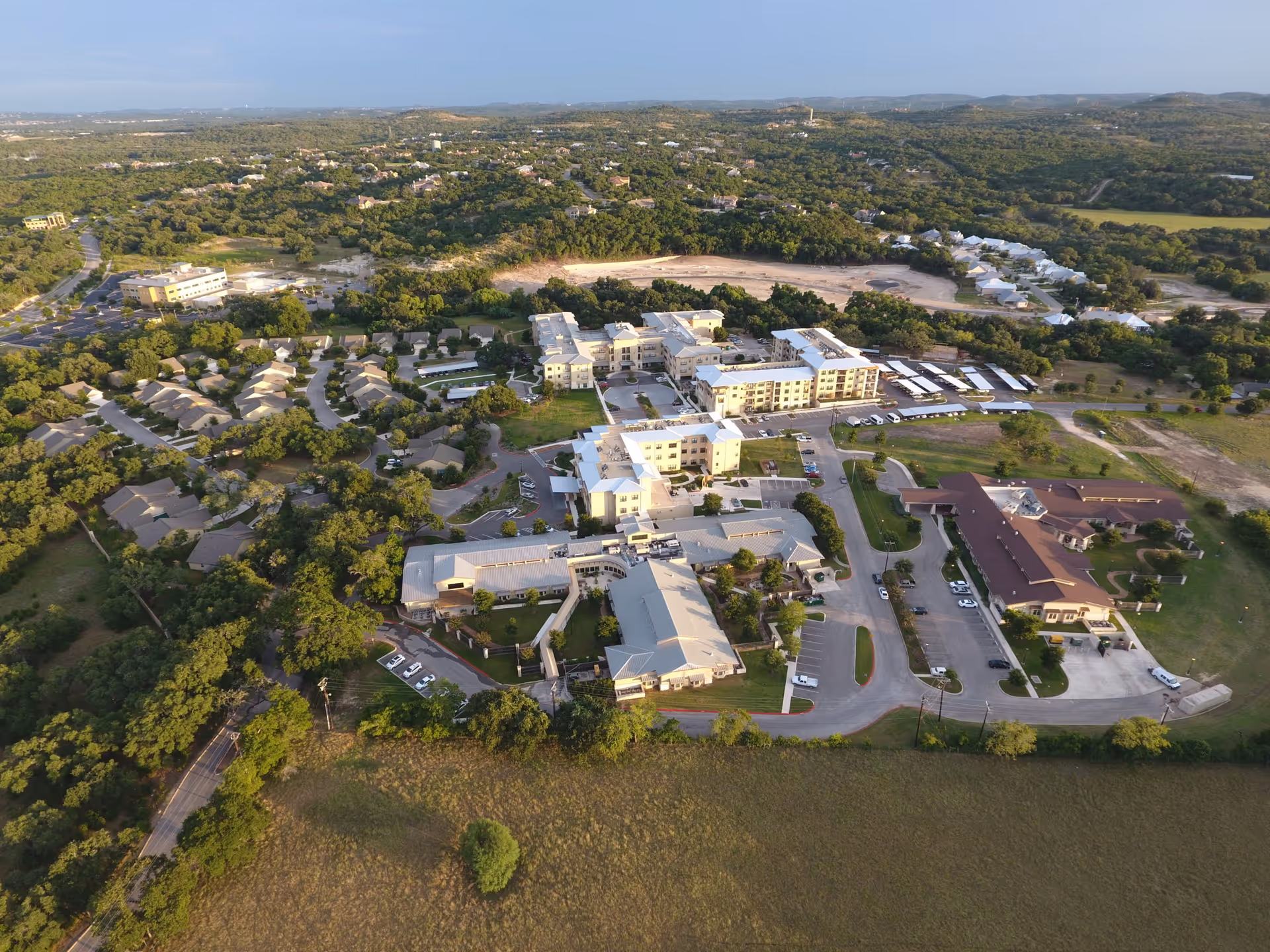 Aerial view of Morningside at Menger Springs - Cibolo House Assisted Living & Memory Care facility surrounded by greenery and trees, showing multiple buildings, parking lots, and roads within a rural landscape.