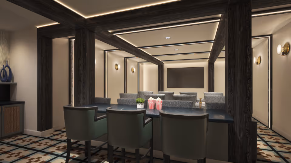 Interior view of a modern common area with a long table surrounded by chairs in the foreground and several armchairs arranged facing a large flat screen TV mounted on the far wall. The room features dark wooden beams, recessed ceiling lighting, wall sconces, and patterned floor tiles.