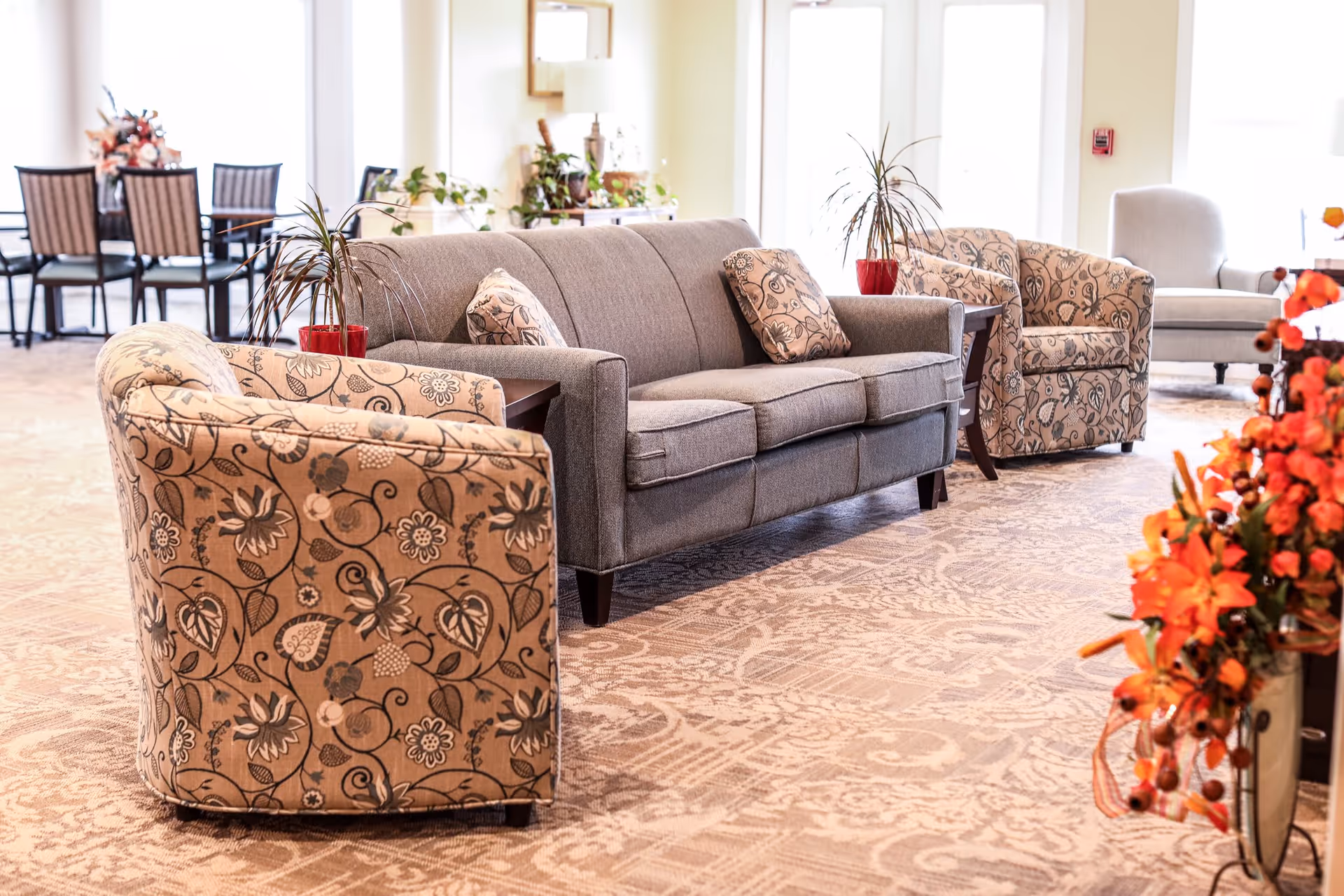 A bright and spacious living room area in an assisted living facility featuring a gray sofa with patterned cushions, two floral-patterned armchairs, a light gray armchair, side tables with potted plants, and a dining table with chairs in the background. The room is decorated with plants and orange flowers, and has large windows allowing natural light to fill the space.